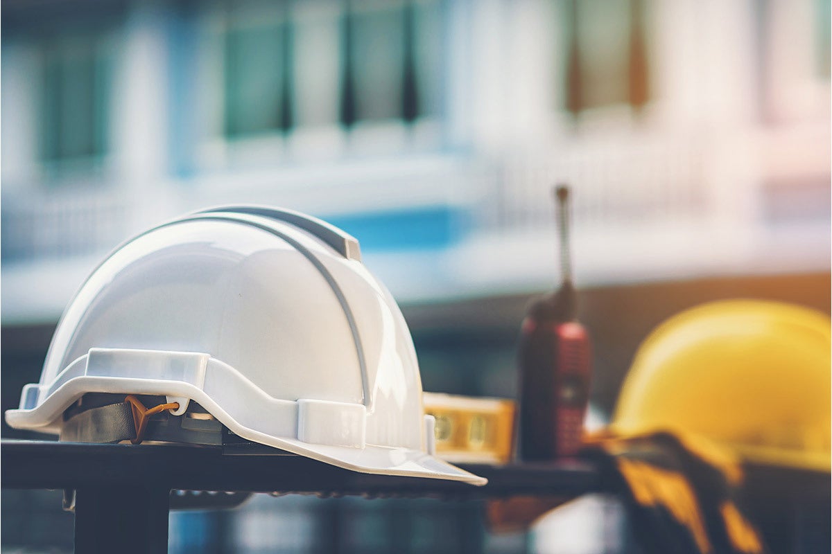 in foreground white and yellow construction hat and walkie-talkie; commercial building blurred in background