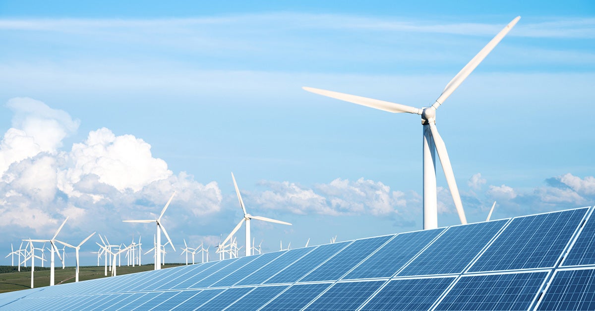 A landscape featuring multiple wind turbines and solar panels under a clear blue sky with scattered clouds. The wind turbines are spread across a green field, and the solar panels are positioned in the foreground, capturing sunlight for renewable energy.