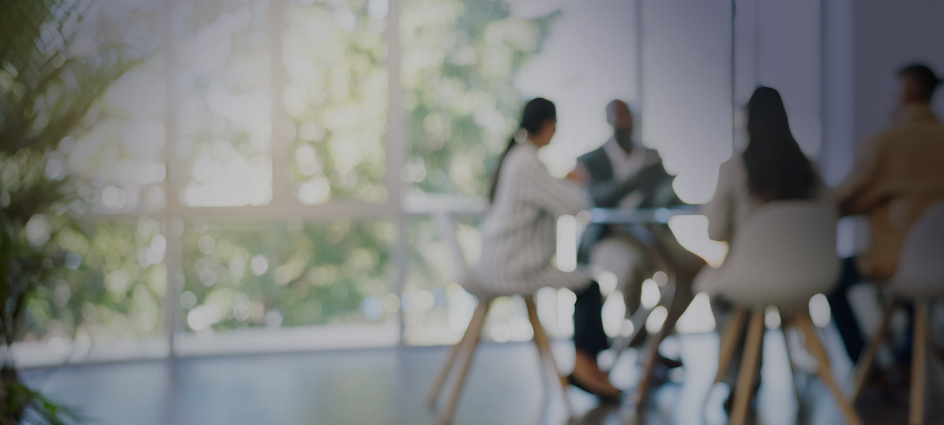 A group of people sitting around a table in a modern office setting. The room is bright with large windows allowing natural light to filter in, creating a soft and inviting atmosphere. The individuals are engaged in conversation, suggesting a collaborative or meeting environment.