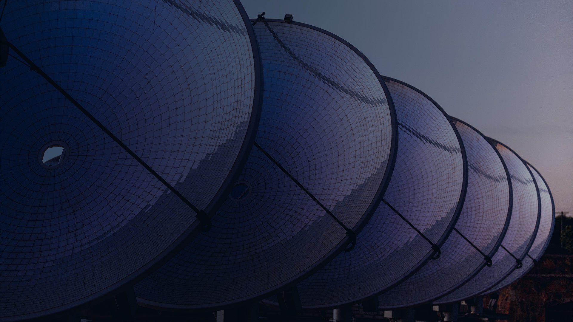 a series of large satellite dishes lined up in a row, facing the same direction. The dishes are composed of a grid-like pattern and appear to be part of a communication or research facility. The sky in the background is dimly lit, suggesting either dawn or dusk. The overall tone of the image is dark, with the dishes reflecting a subtle blue hue.