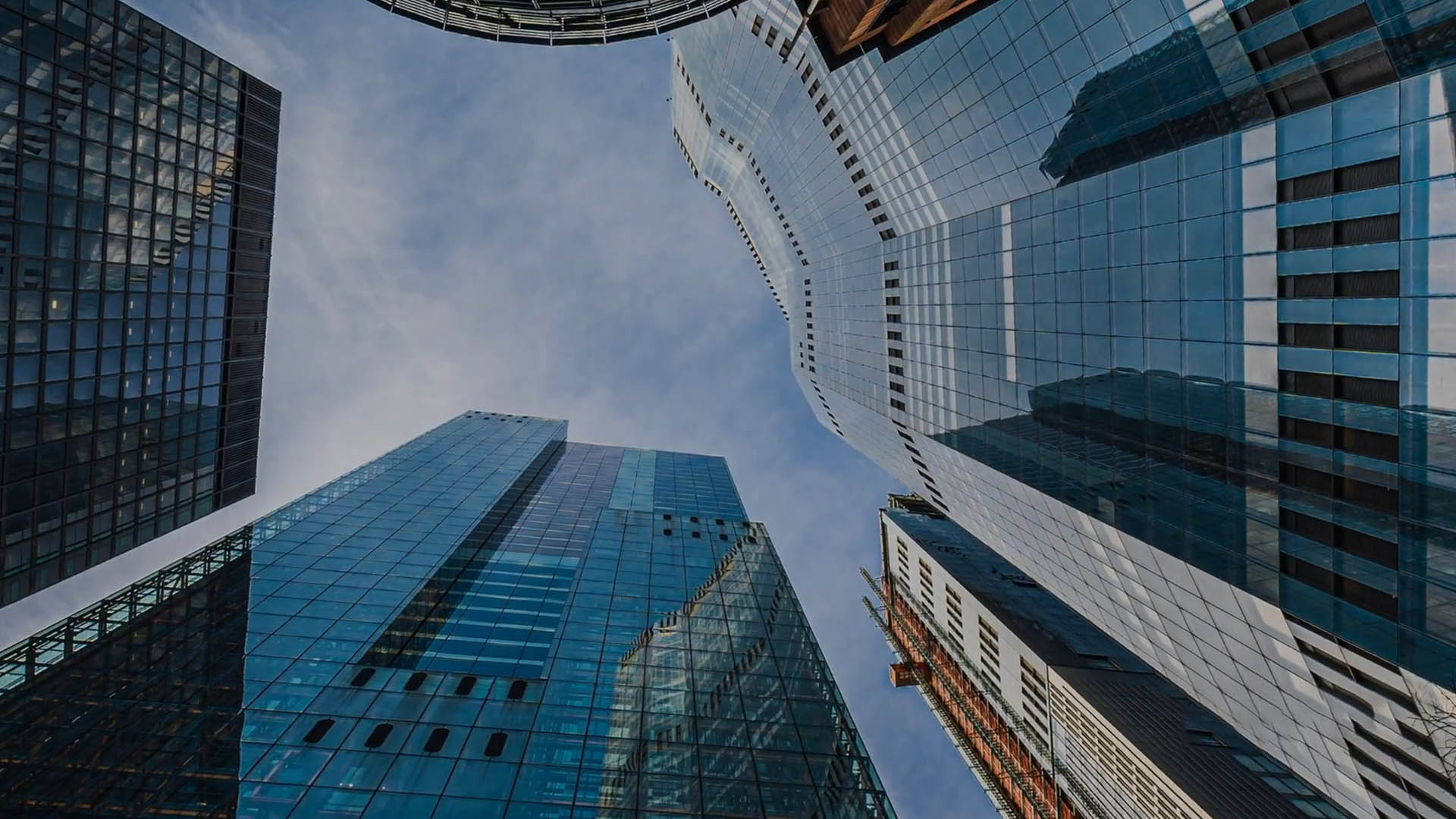 Upward view of several modern glass skyscrapers against a blue sky, with reflections on the buildings' surfaces.
