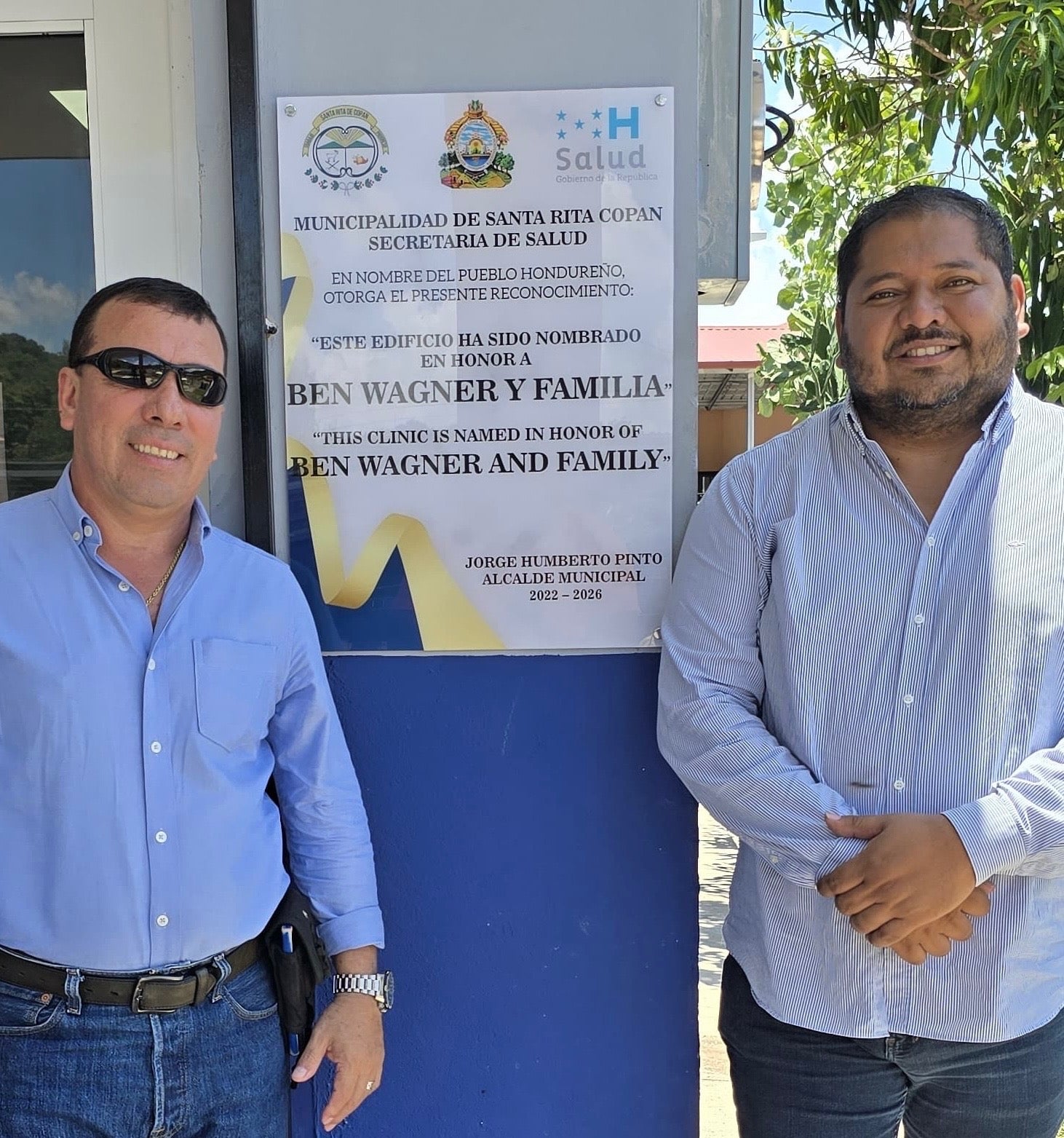 Two men stand in front of a commemorative plaque mounted on a blue wall outside a building. The plaque features the logos of the Santa Rita Copán Municipality and the Health Department of Honduras, and is written in both Spanish and English. It states that the building is named in honor of "Ben Wagner and Family," and is signed by Jorge Humberto Pinto, Municipal Mayor, for the term 2022-2026. The men are wearing long-sleeve button-up shirts and jeans, and the scene is outdoors with trees and a building visible in the background.