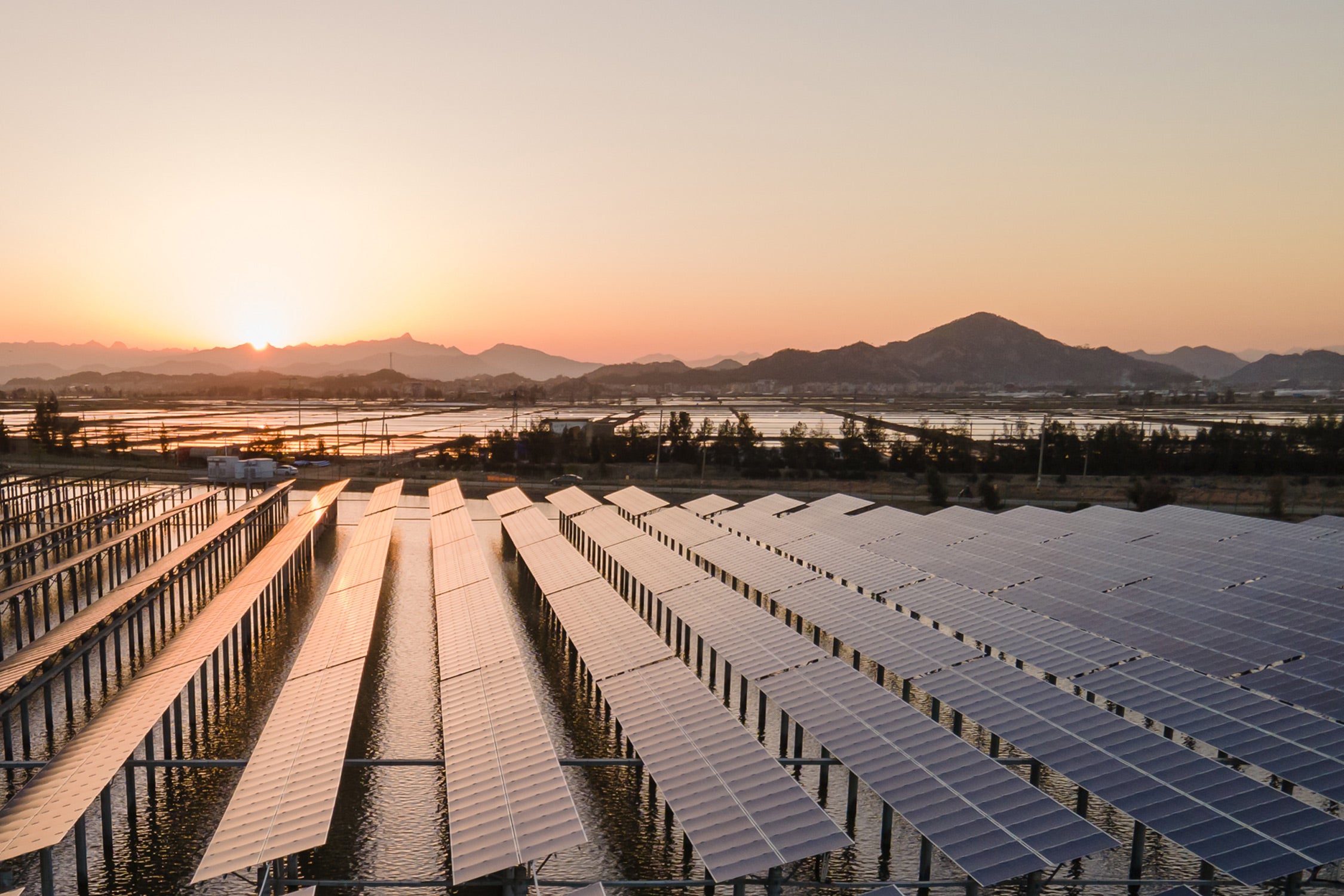 Rows of solar panels installed above water, reflecting the sunlight at sunset, with mountains and trees in the background.