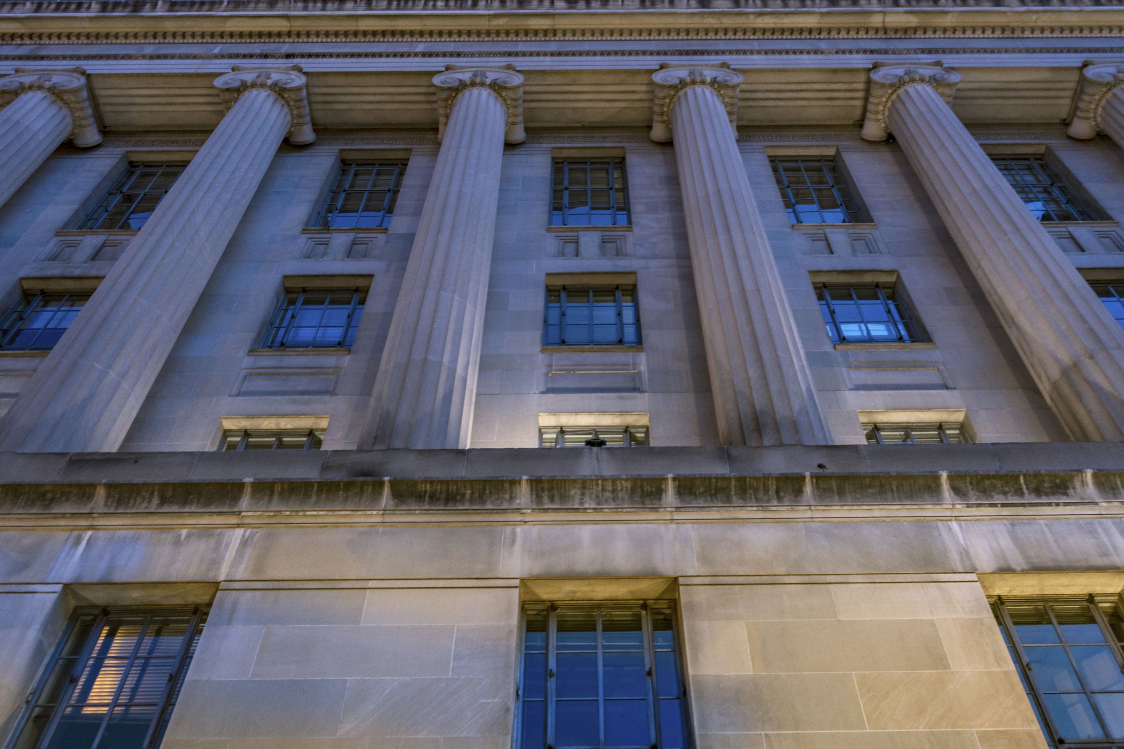 Upward view of a tall neoclassical stone building at dusk, with fluted columns and rectangular windows, lit by cool blue evening light and warm spotlights that emphasize the building’s height and symmetry.