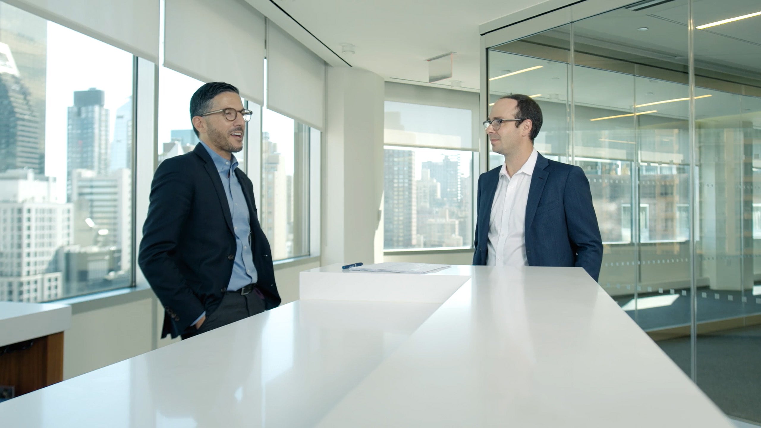 Two business professionals in suits talking at a white conference table in a modern law office with floor-to-ceiling windows and a city skyline in the background